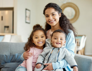 A mother sitting on the couch with her toddler daughter and infant son smiling for a photo